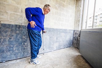 Senior woman standing at a wall with a crutch, having severe hip pain.